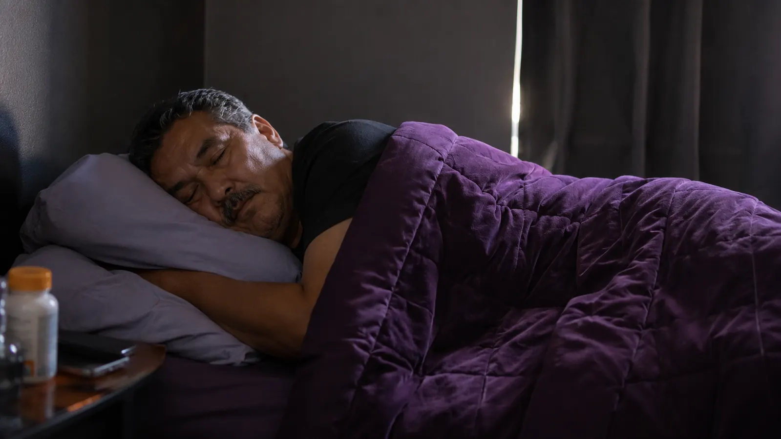 Middle-aged man sleeping soundly on his side under a purple weighted blanket in a dark bedroom