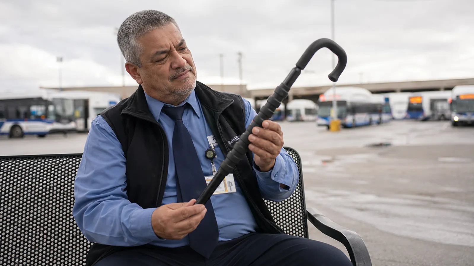 Middle-aged bus driver sitting on a bench outside a transit depot, holding a Body Back Buddy massage cane and looking skeptically at the packaging