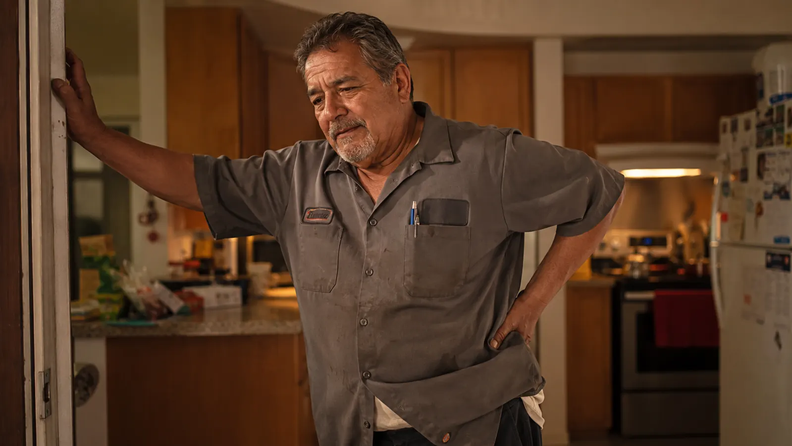 Man in his fifties in work clothes stretching his lower back in a kitchen doorway after an evening shift