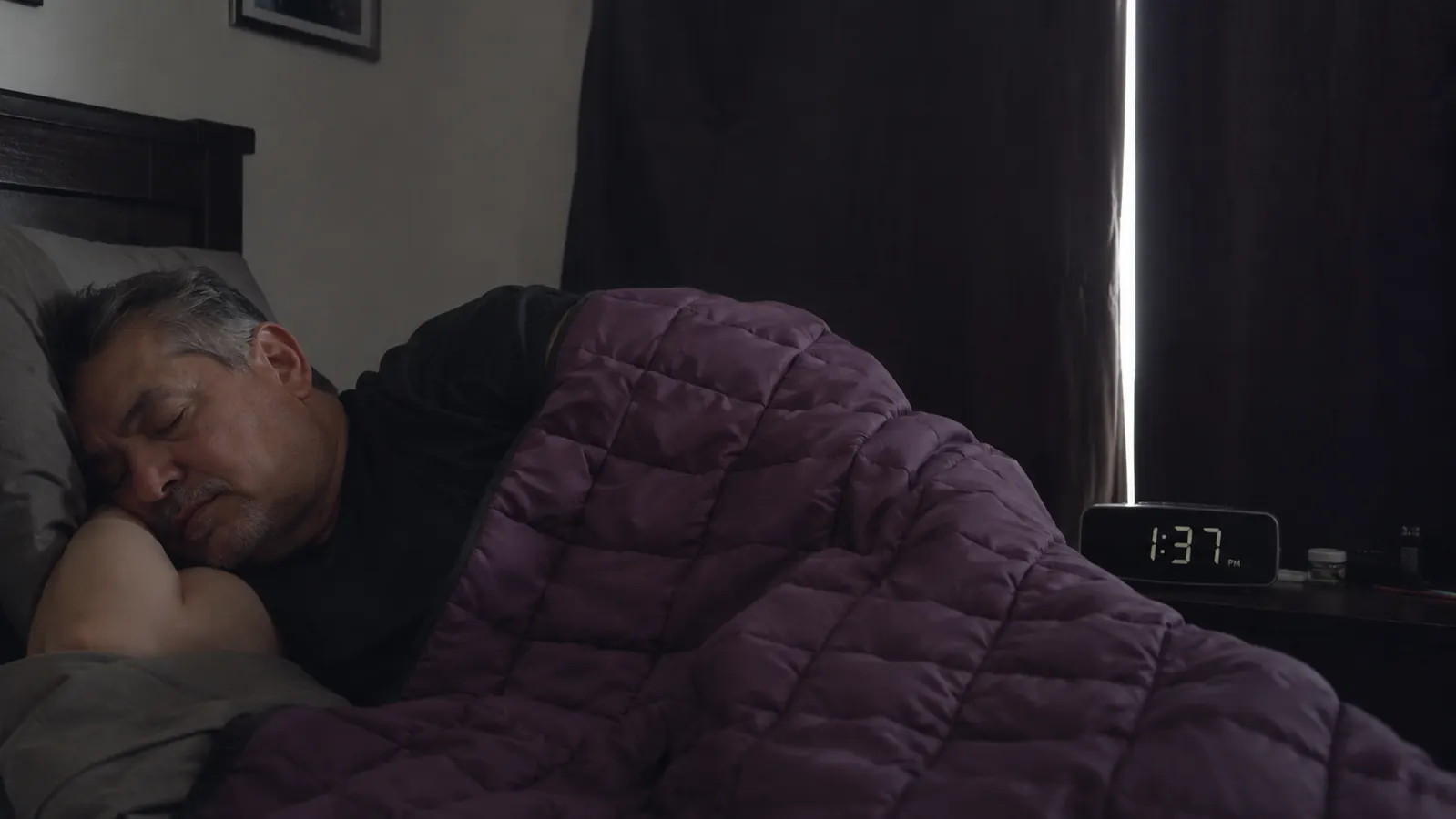 Middle-aged man sleeping on his side under a heavy blanket in a dim bedroom with blackout curtains drawn, midday light barely leaking around the edges
