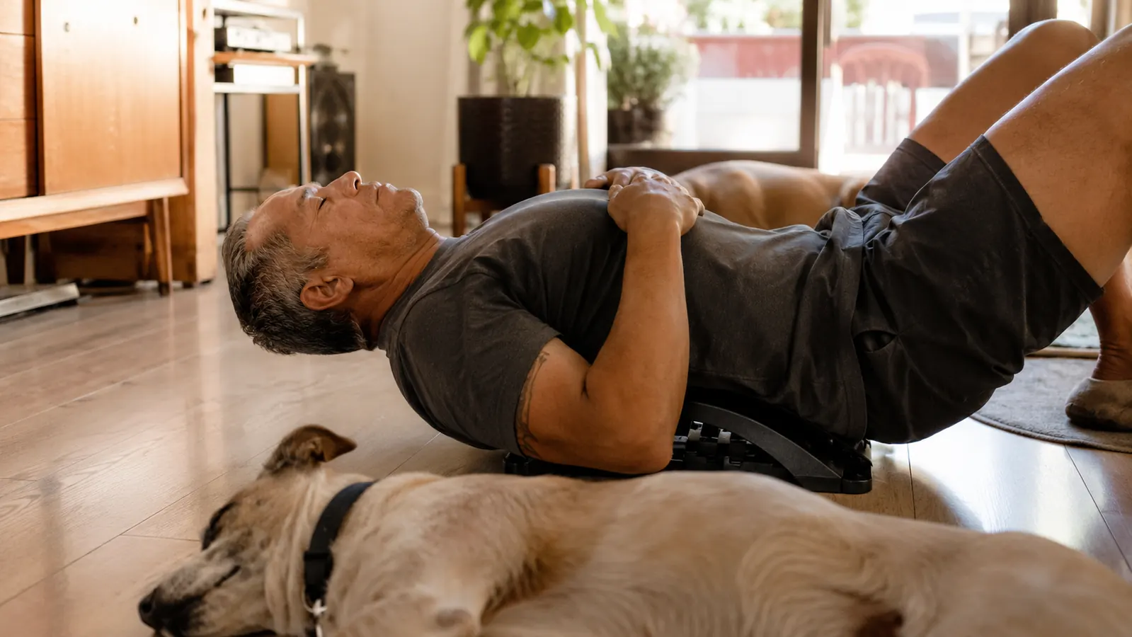 Middle-aged man in pajama pants lying on a living room floor with his lower back arched over an adjustable back stretcher arc in the early morning light