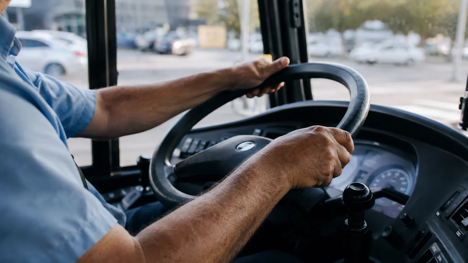 A man's hands gripping a bus steering wheel, knuckles visible, morning light coming through the windshield
