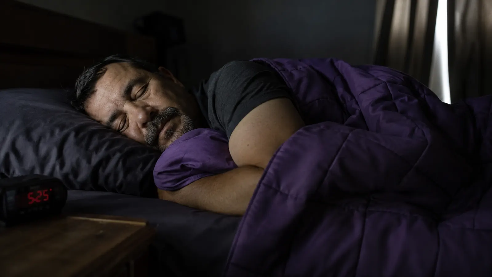 Middle-aged man asleep under a purple weighted blanket in a dim bedroom with morning light at the edge of the curtains