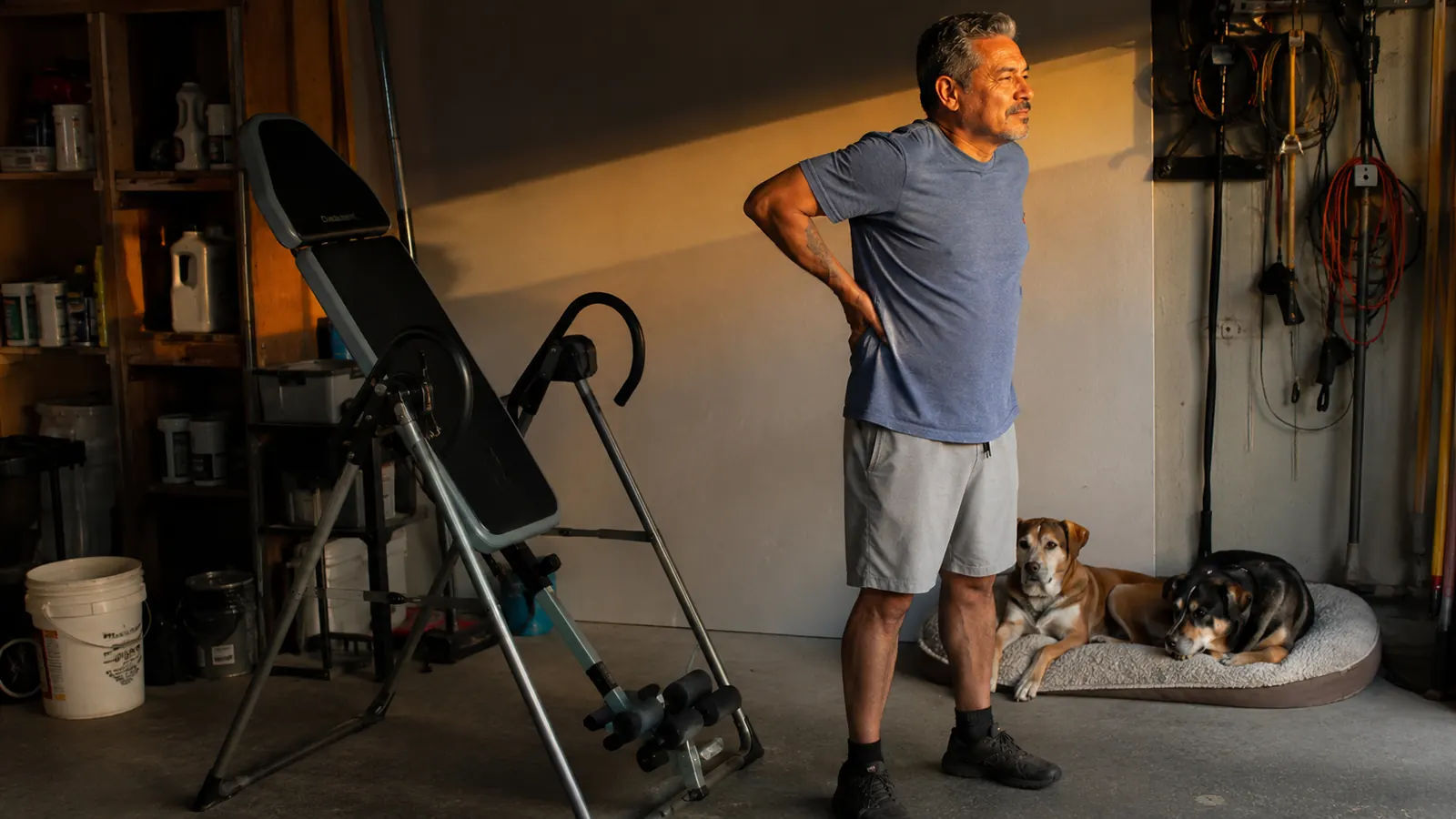Man standing upright and stretching his lower back in a garage after finishing an inversion session, two dogs resting nearby